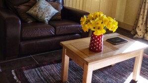 Un salon avec un canapé en cuir marron, une table basse en bois sur laquelle est posé un vase de fleurs jaunes, et une porte visible au fond.