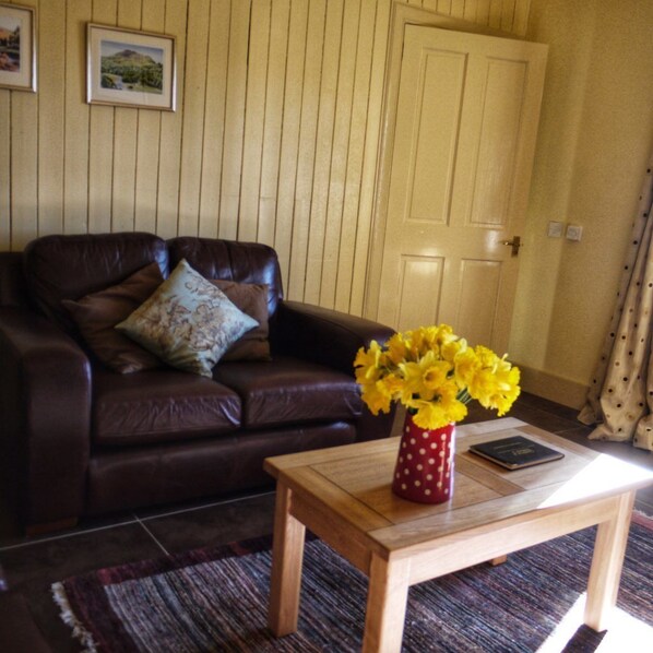 A living room with a brown leather sofa, a wooden coffee table with a vase of yellow flowers, and a door in the background.