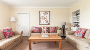A living room with two beige sofas, a wooden coffee table, a framed dog artwork, a white door, and a bookshelf with books. - Garvard (Isle of Colonsay)