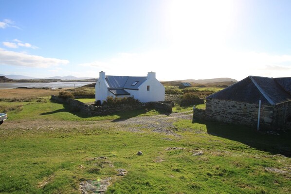 A white house with a sloped roof and solar panels, surrounded by a grassy field and a stone wall.