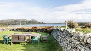 A patio with a table and chairs, a stone wall, and a view of the sea and hills. - Garvard (Isle of Colonsay)