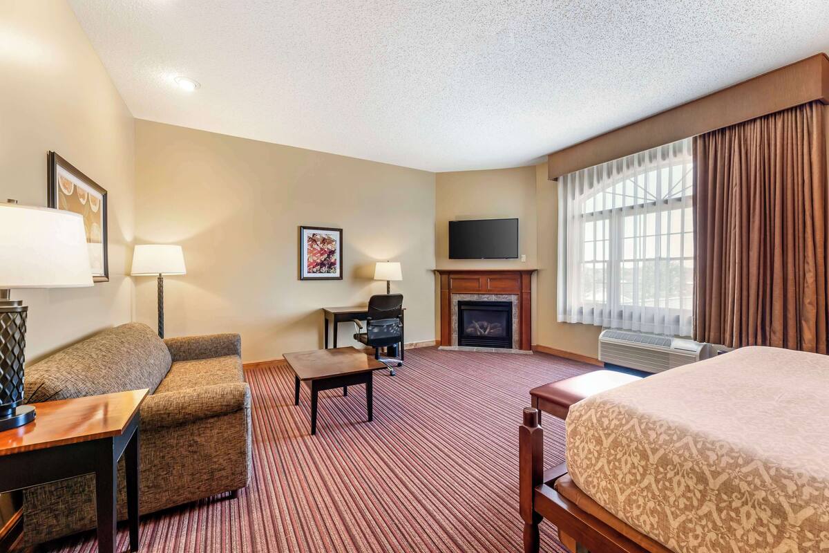 A hotel room with a bed, a sofa, a desk with a lamp, a fireplace, a TV, and a large window with curtains.