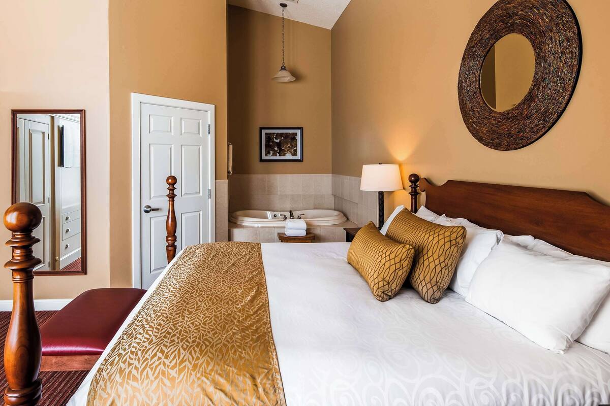 A hotel room with a large bed, a wooden headboard, a round mirror, a bathroom with a bathtub, and a red bench.