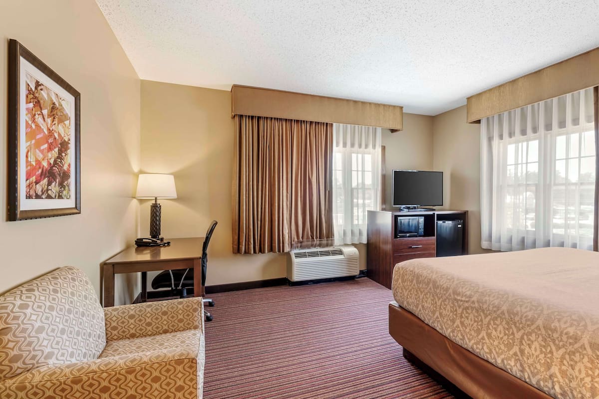 A hotel room with a bed, a desk with a lamp, a television, and a large window with curtains.