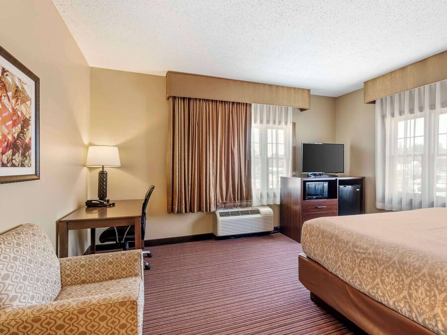 A hotel room with a bed, a desk with a lamp, a television, and a large window with curtains.