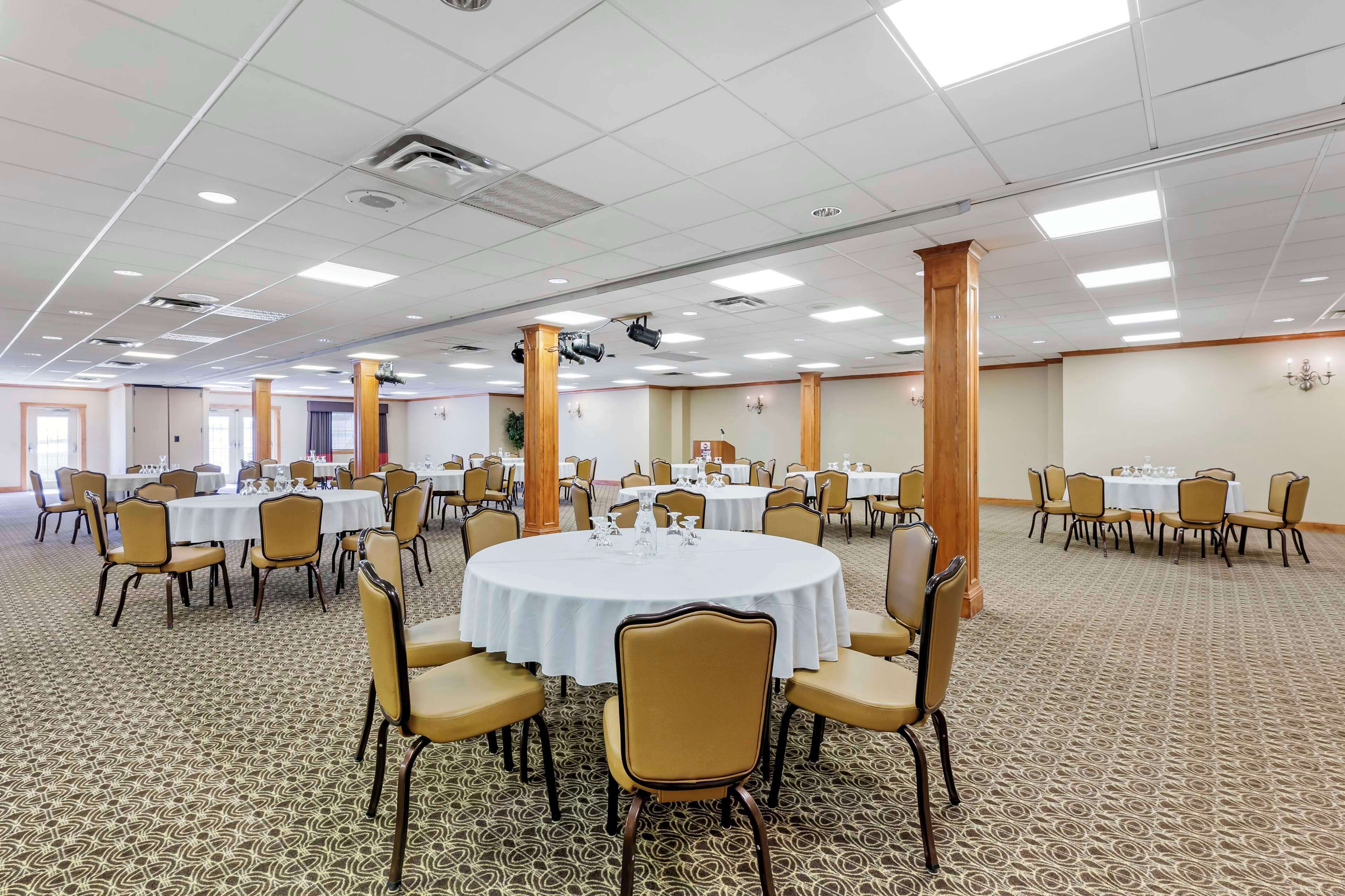 A large, well-lit banquet hall with multiple round tables, each surrounded by chairs. The room has a patterned carpet and a high ceiling with recessed lighting.