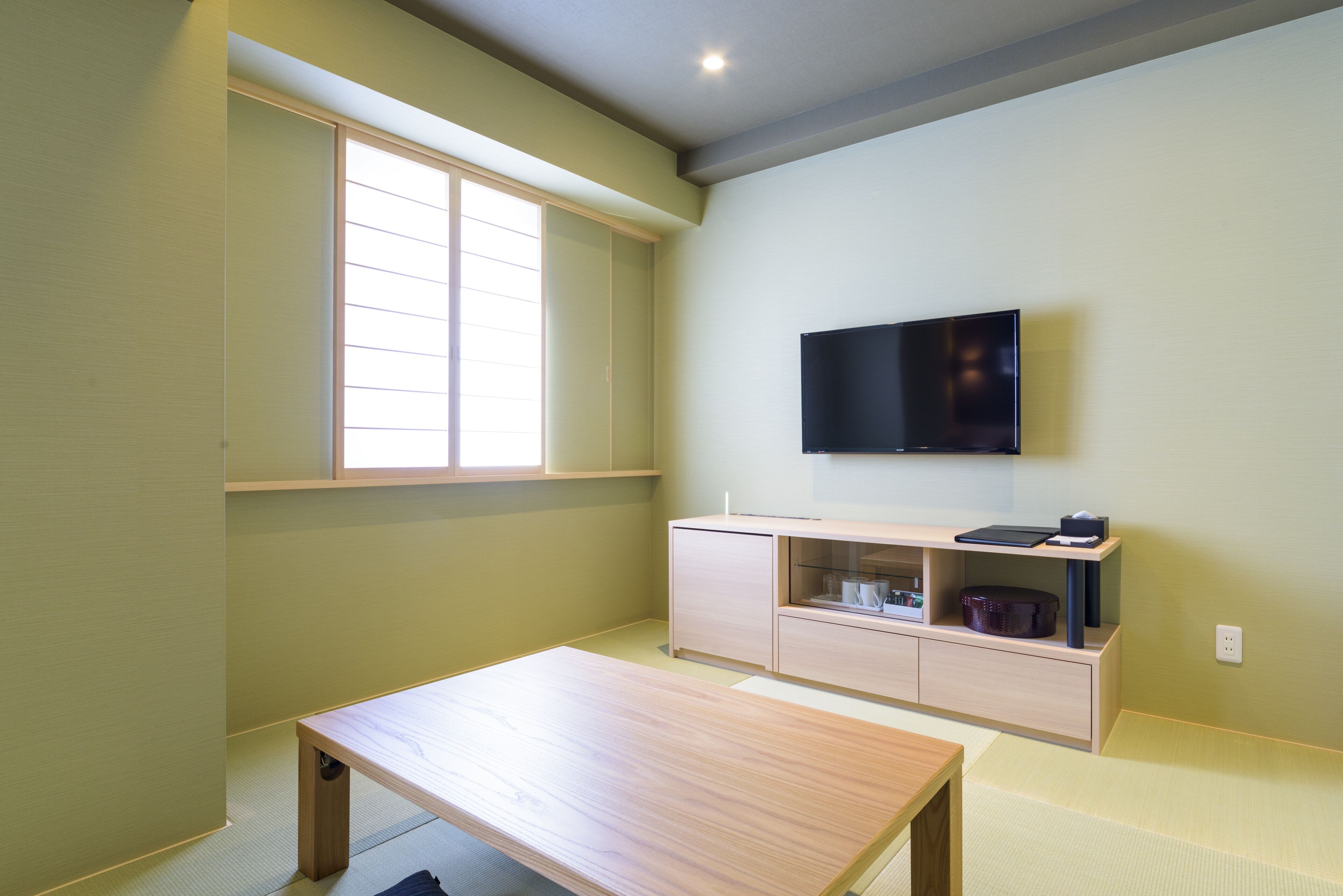 Una habitación con una mesa de madera, un televisor montado en la pared, una ventana con persianas y un piso de color claro.