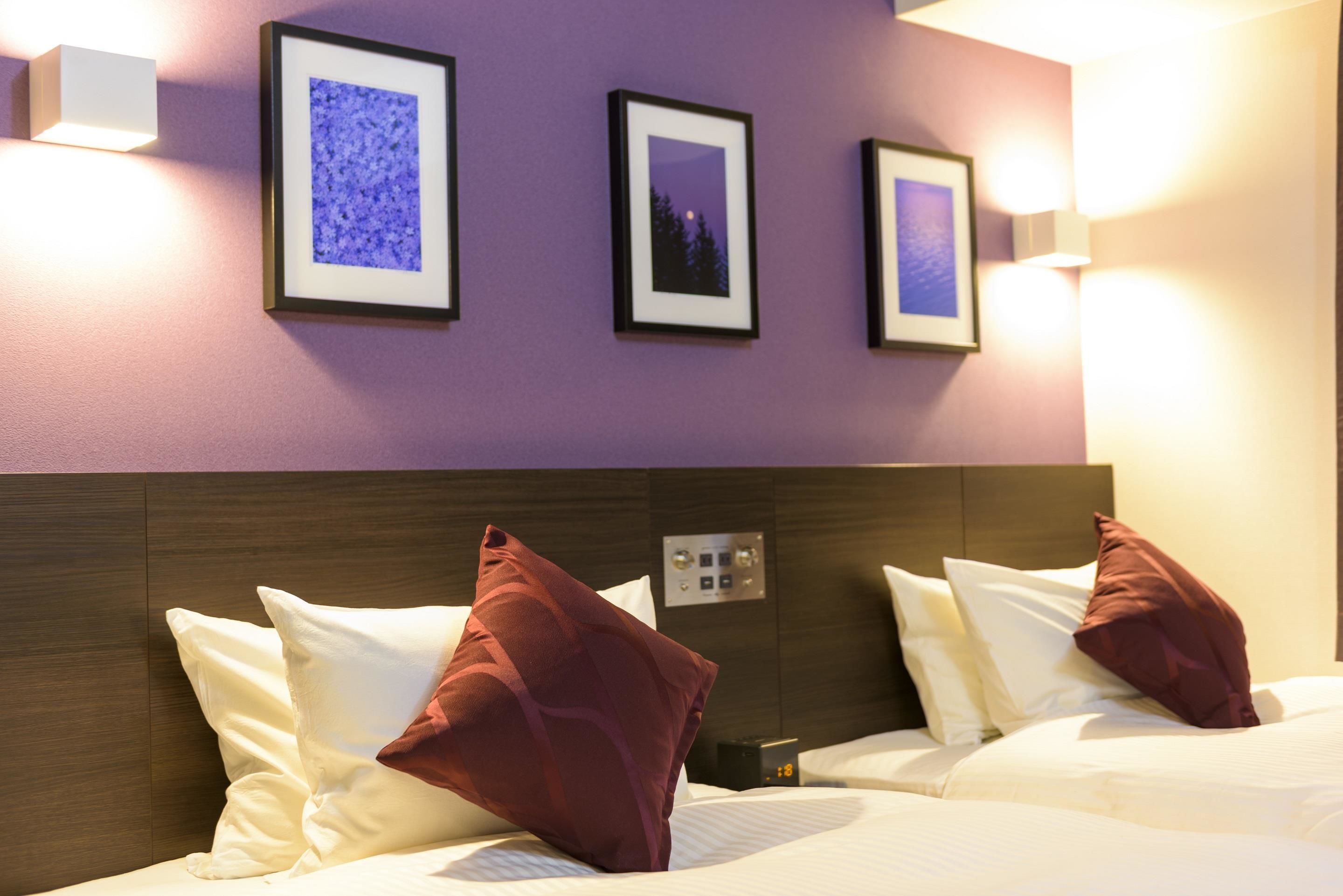 A hotel room with two beds, each with white bedding and red pillows, a wooden headboard, and three framed pictures on the wall.