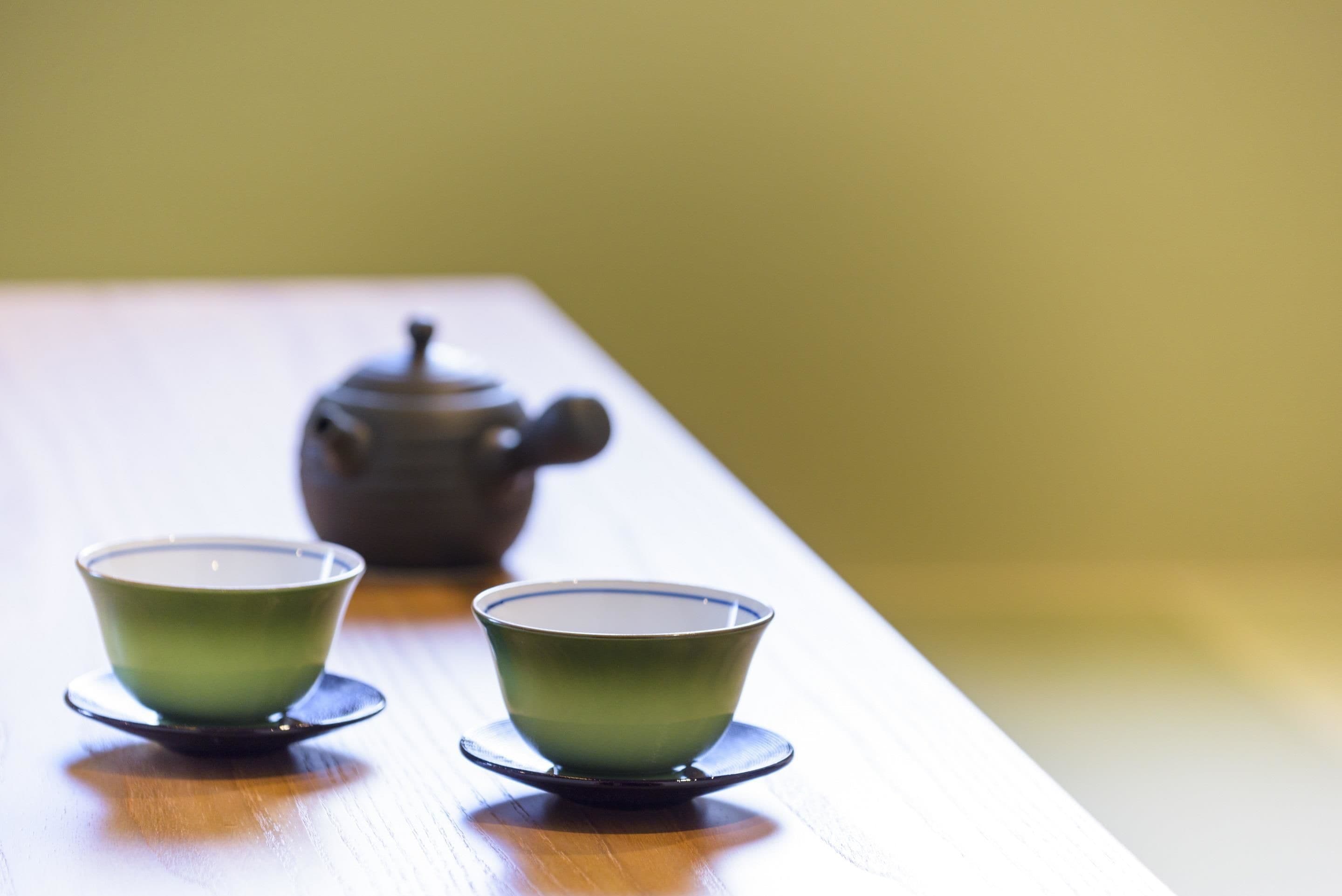 Two green cups on a wooden table with a teapot in the background.