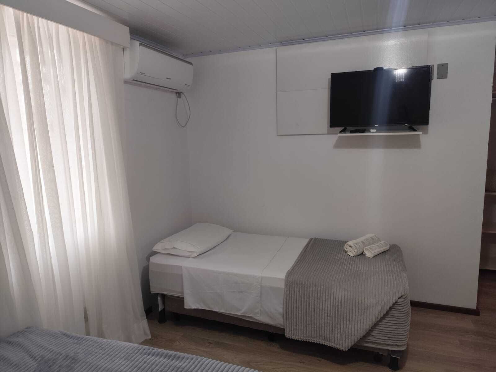 Una cama individual con colchón blanco y manta gris, un televisor montado en la pared y una ventana con cortinas blancas.