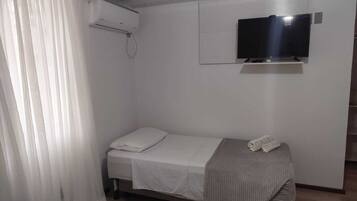 Una cama individual con colchón blanco y manta gris, un televisor montado en la pared y una ventana con cortinas blancas.