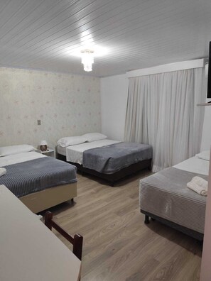 A hotel room with three beds, a wooden floor, a flat-screen TV, and a window with curtains.