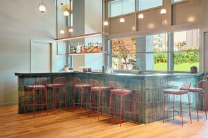 A modern bar area with a wooden floor, red bar stools, and a large window offering a view of greenery.
