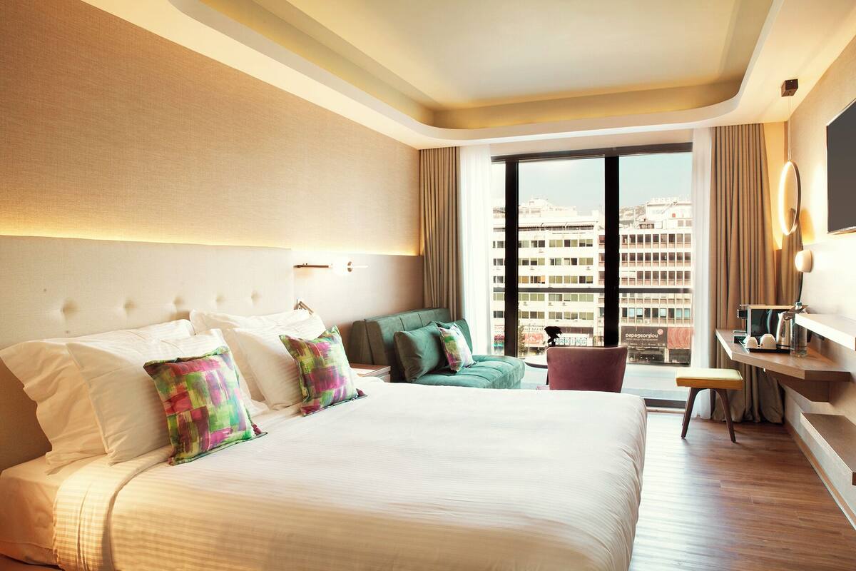 A hotel room with a large bed, a sofa, a desk, and a view of the city through large windows.