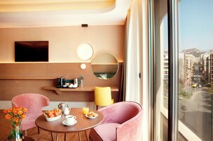 Quarto de hotel moderno com uma grande janela, um espelho redondo, televisão de tela plana, espaço para café e duas cadeiras cor de rosa.