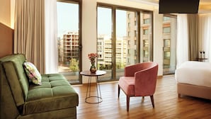 A modern hotel room with a green sofa, a pink armchair, and a white bed. Large windows offer a view of the cityscape. - Zeus Essence Wyndham Athens Residence (Athens)