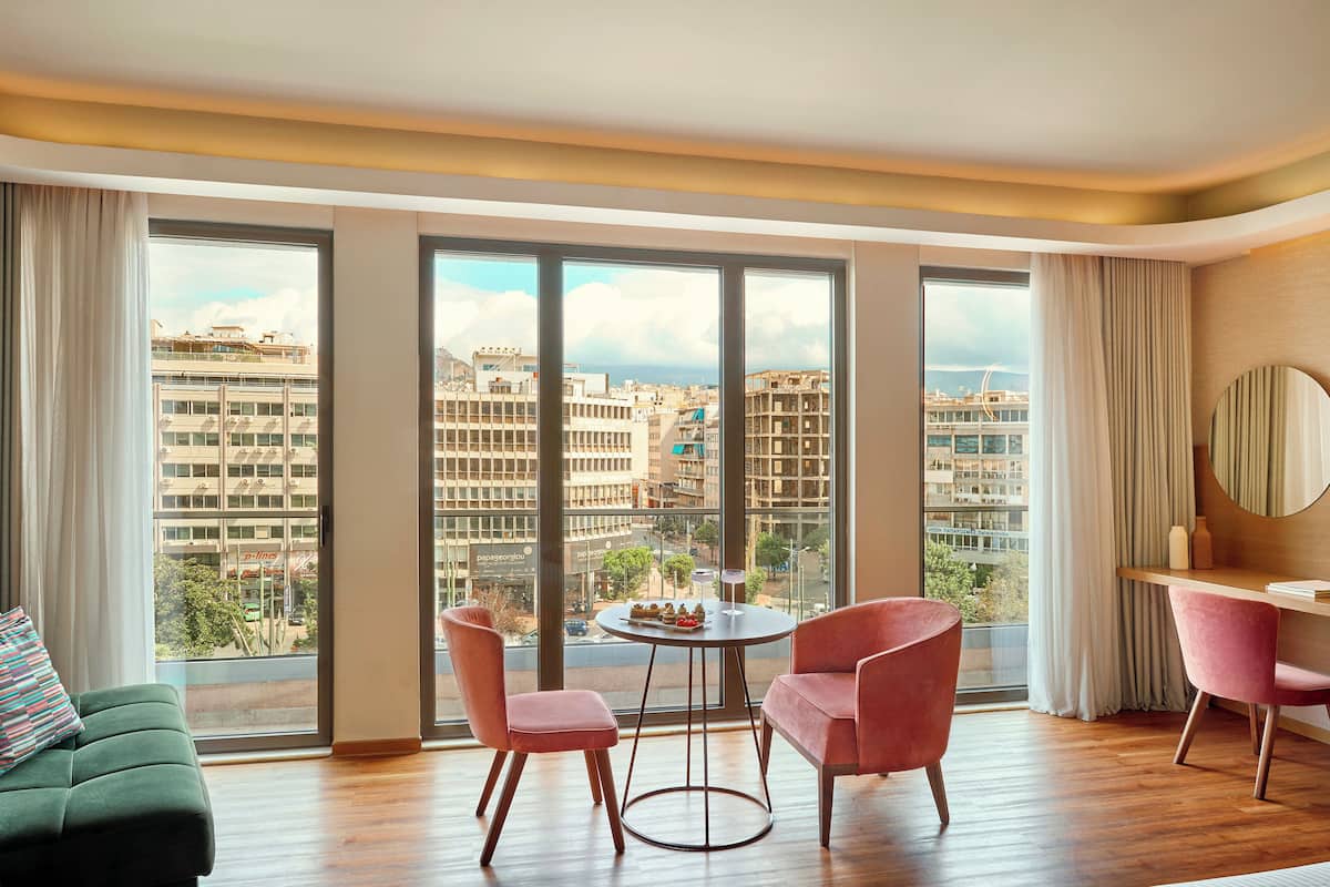 A modern hotel room with large windows, a round table, two chairs, a sofa, and a desk with a mirror.