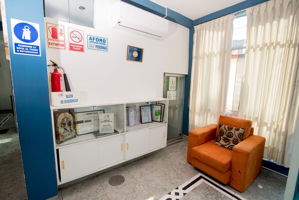 Una zona de recepción con decoración azul y blanca, un extintor, un cartel de "Prohibido Fumar", un cartel que indica "Cúbrete la boca al toser o estornudar", un cartel de "AFORO" y un certificado enmarcado en la pared.