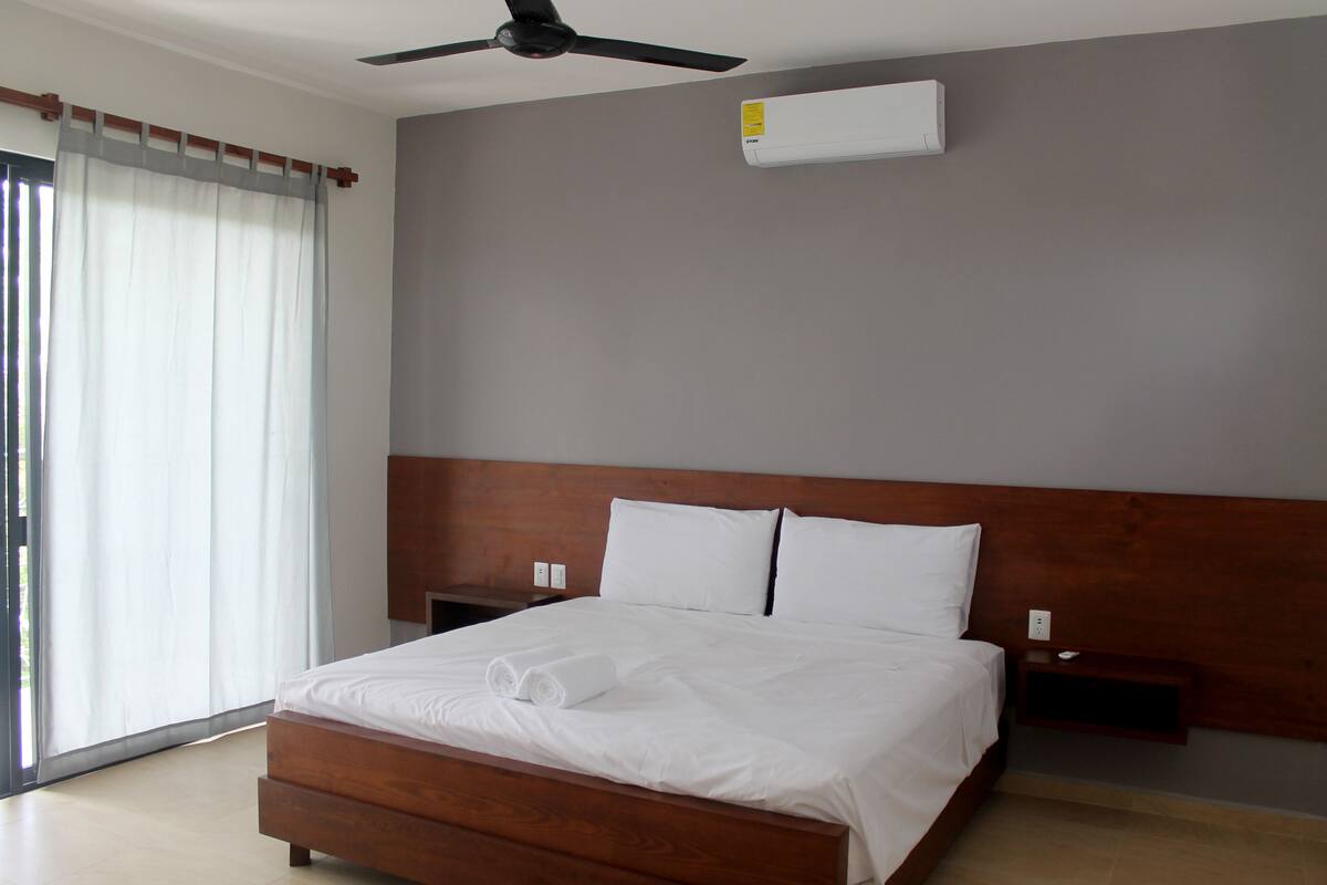 A modern hotel room with a large bed, wooden headboard, ceiling fan, air conditioning unit, and a nightstand.