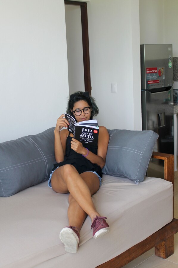 A person sitting on a grey sofa with a book titled "Roberta Como Artista" in a room with a white wall and a stainless steel appliance in the background.