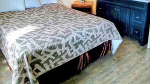 Un lit bien fait, recouvert d’une couvre-lit à motifs, une lampe de chevet, une commode de couleur sombre avec un miroir, et un petit vase avec des fleurs posé sur la commode.