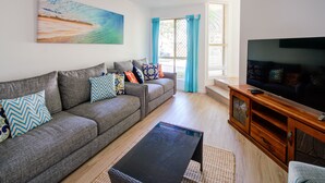 Un salon avec un canapé gris, une table basse noire, un meuble TV en bois et un tableau représentant une scène de plage.
