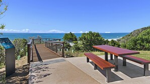 Une terrasse en bois menant à une plage, avec des tables de pique-nique et un panneau indicateur.
