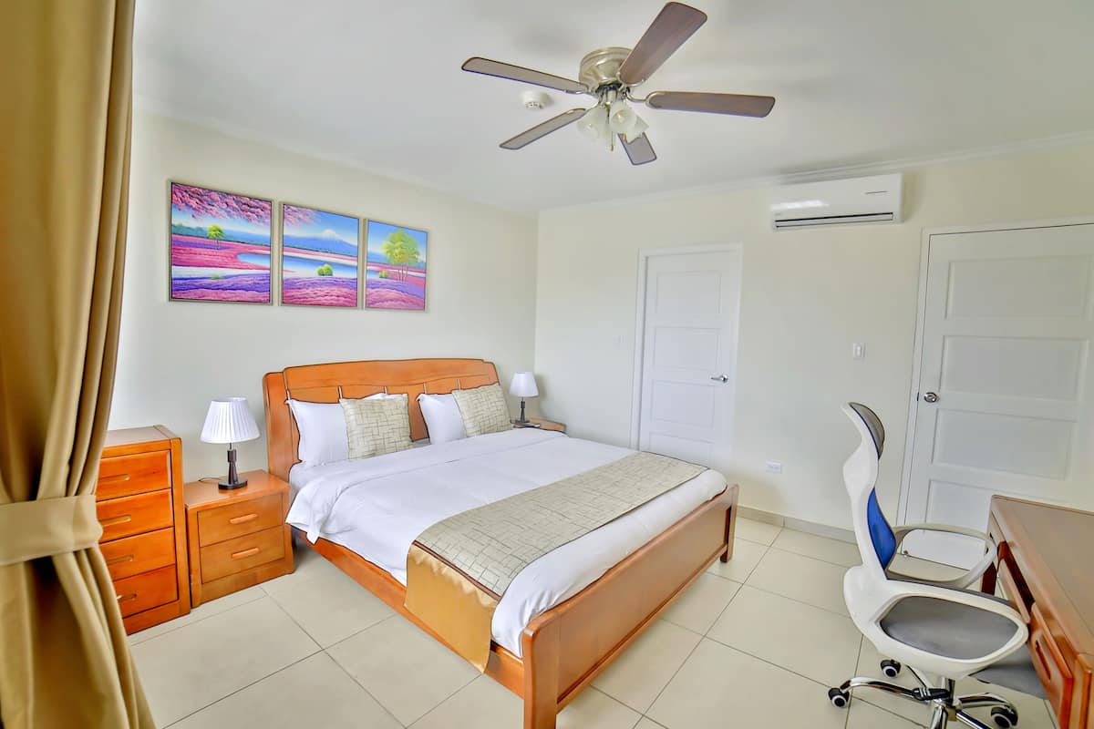 Quarto com cama de madeira, mesa de cabeceira, escrivaninha com cadeira e ventoinha no teto.