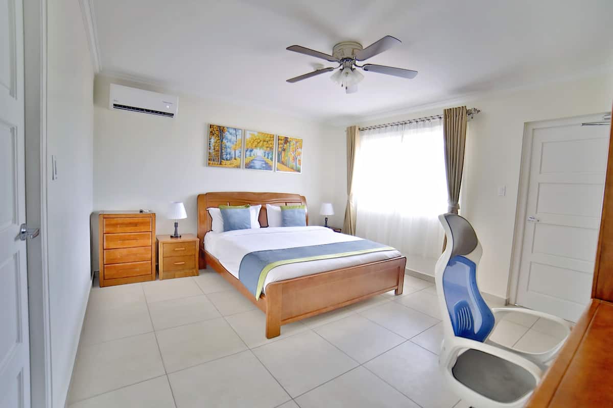 Quarto com cama, cómoda de madeira, mesa de cabeceira, ventoinha no teto e janela com cortinas.