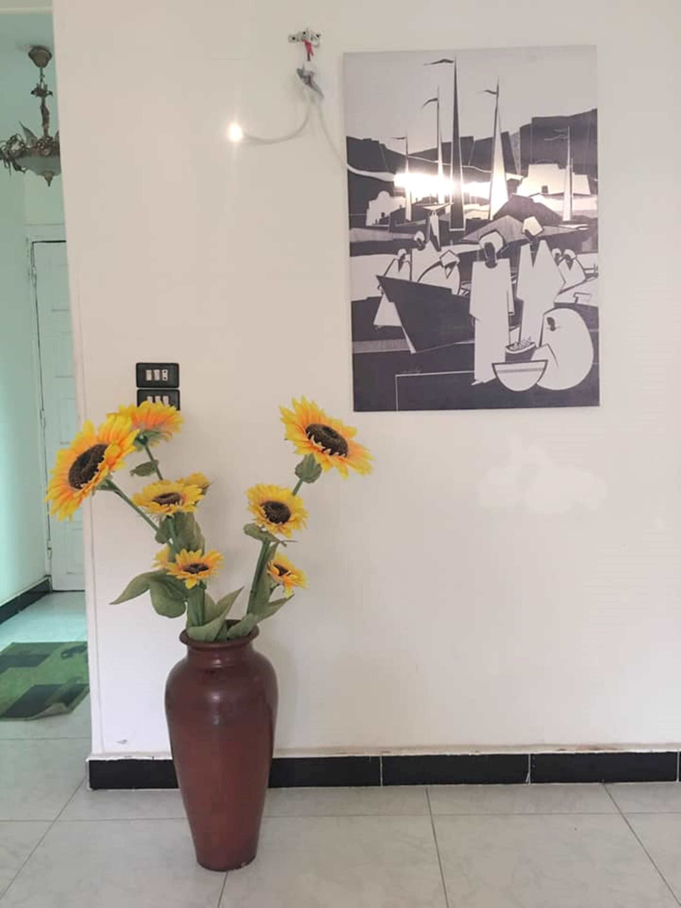 Uma parede com uma pintura a preto e branco de uma cena portuária, um vaso com girassóis e uma porta com um letreiro.