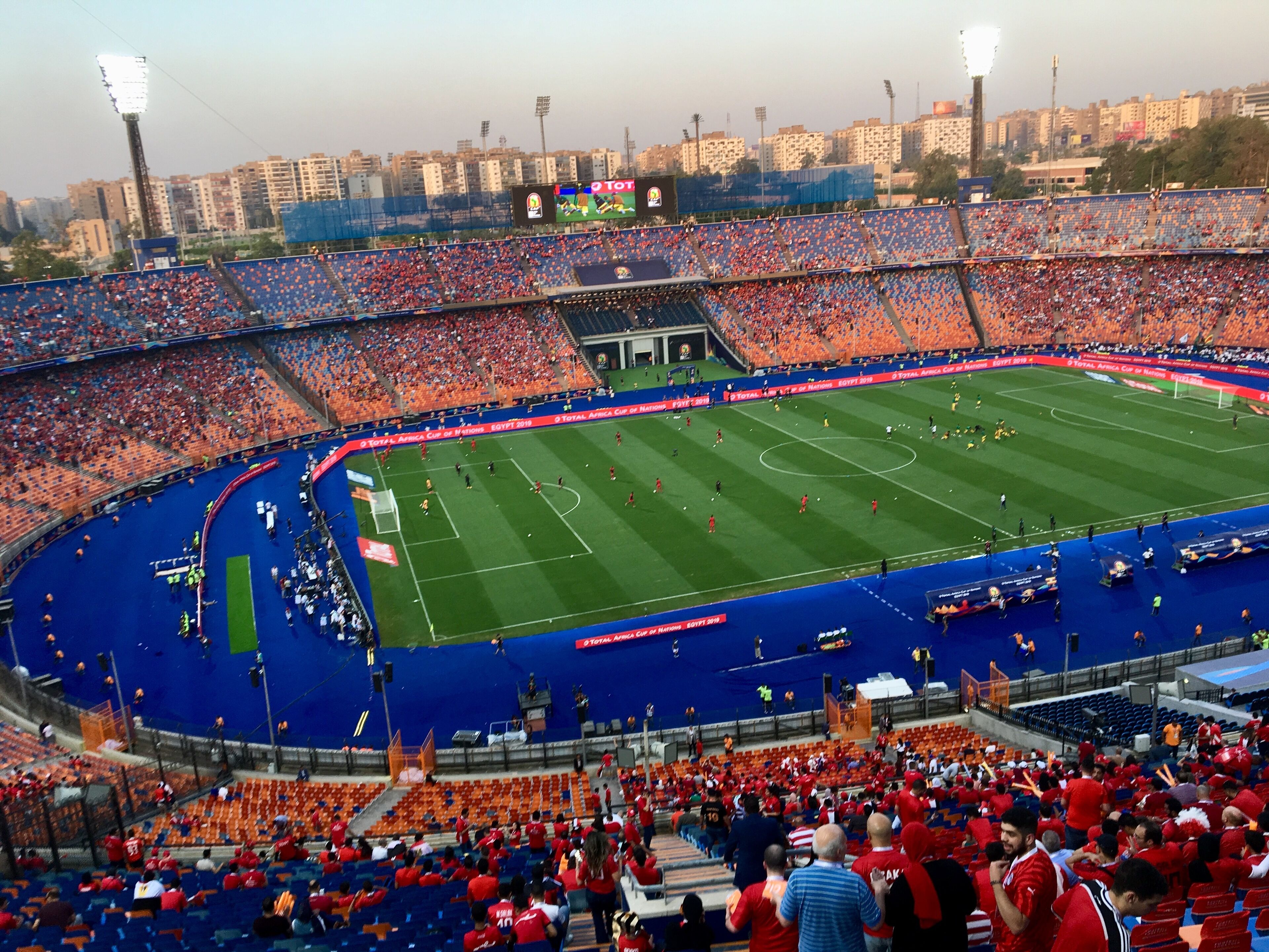 A stadium with multiple tiers of seating, a central field, and surrounding urban buildings.
