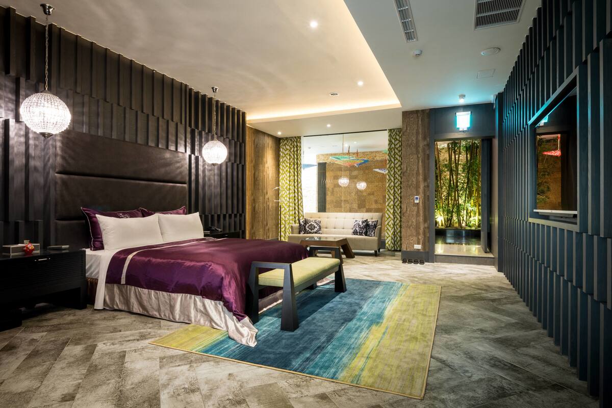 A modern bedroom with a large bed, bedside tables, a bench, and a view of a lush garden through a glass door.
