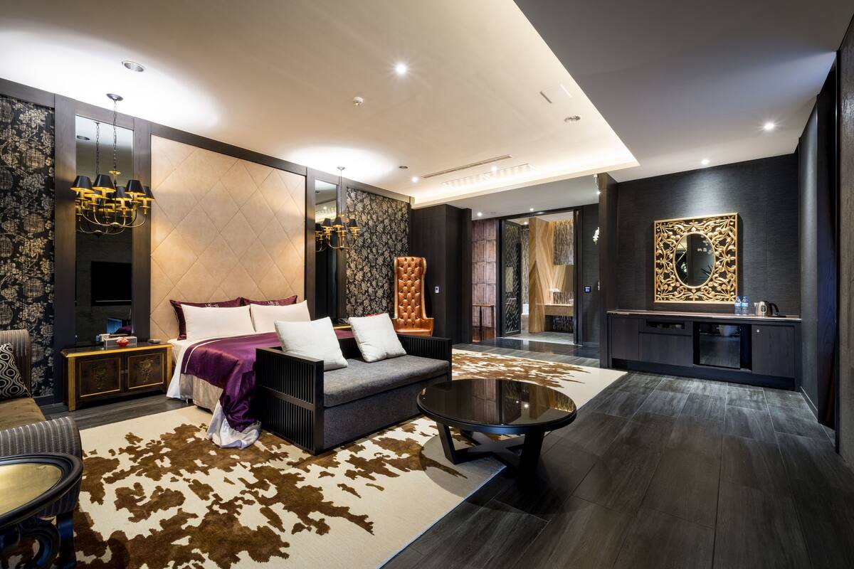 A modern hotel room with a large bed, a sofa, a coffee table, and a dining area with a dark wood finish.
