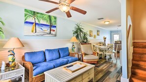A living room with a blue sofa, a ceiling fan, a painting of a beach scene, and a dining area with chairs and a table. - Near Port Aransas Beach: Condo w/ Pool Access (Port Aransas)