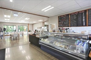 A cafeteria with a glass display case filled with food items, a chalkboard menu, and a seating area with tables and chairs. - Tui Base Camp (Tuatapere)