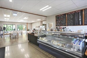 Una cafetería con un mostrador de vidrio lleno de alimentos, un menú en la pizarra y una zona de estar con mesas y sillas.