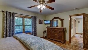 Une chambre à coucher comprenant un lit, une commode en bois, un ventilateur de plafond, un téléviseur et une fenêtre avec des rideaux.
