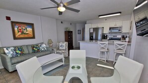 A modern living room with a glass dining table, white chairs, a sofa with colorful cushions, a ceiling fan, and a kitchen area with white cabinets and appliances. - Beachfront Condo w/ Indoor Outdoor Pool & Grills | Gulf Shores | Sleeps 6 (Orange Beach)
