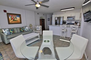 A modern living room with a glass dining table, white chairs, a sofa with colorful cushions, a ceiling fan, and a kitchen area with white cabinets and appliances.