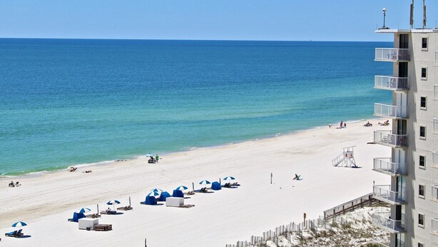 A beach with white sand, clear blue water, and a building with balconies overlooking the beach. - Beachfront Condo w/ Indoor Outdoor Pool & Grills | Gulf Shores | Sleeps 6 (Orange Beach)