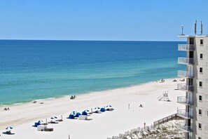 A beach with white sand, clear blue water, and a building with balconies overlooking the beach.