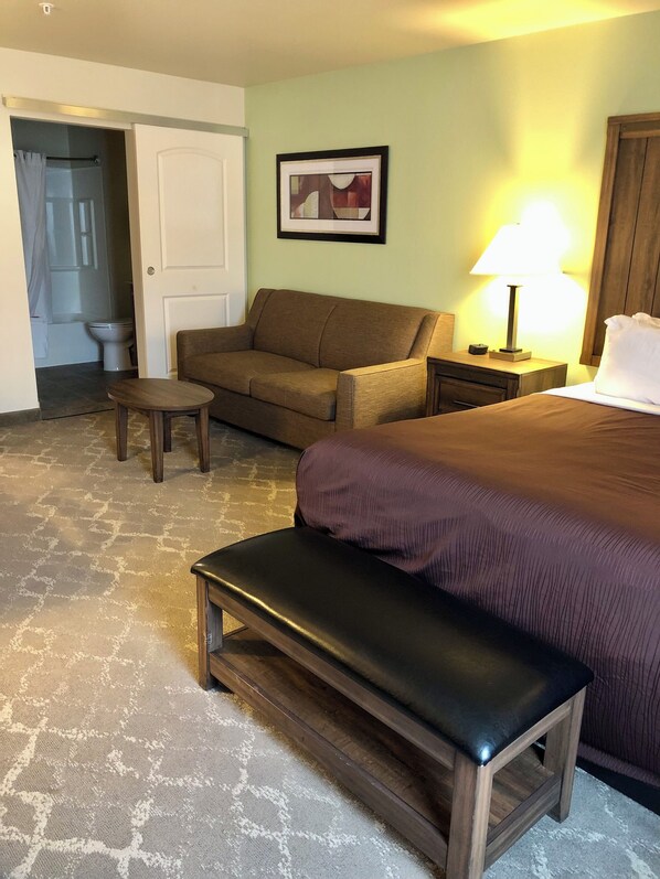 Habitación de hotel con cama, sofá, una mesita, un banco, una mesita de noche con lámpara, un cuadro en la pared y una puerta que da a un baño.