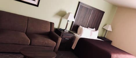 A hotel room with a brown sofa, a small wooden table, a bed with a headboard, and two bedside lamps.
