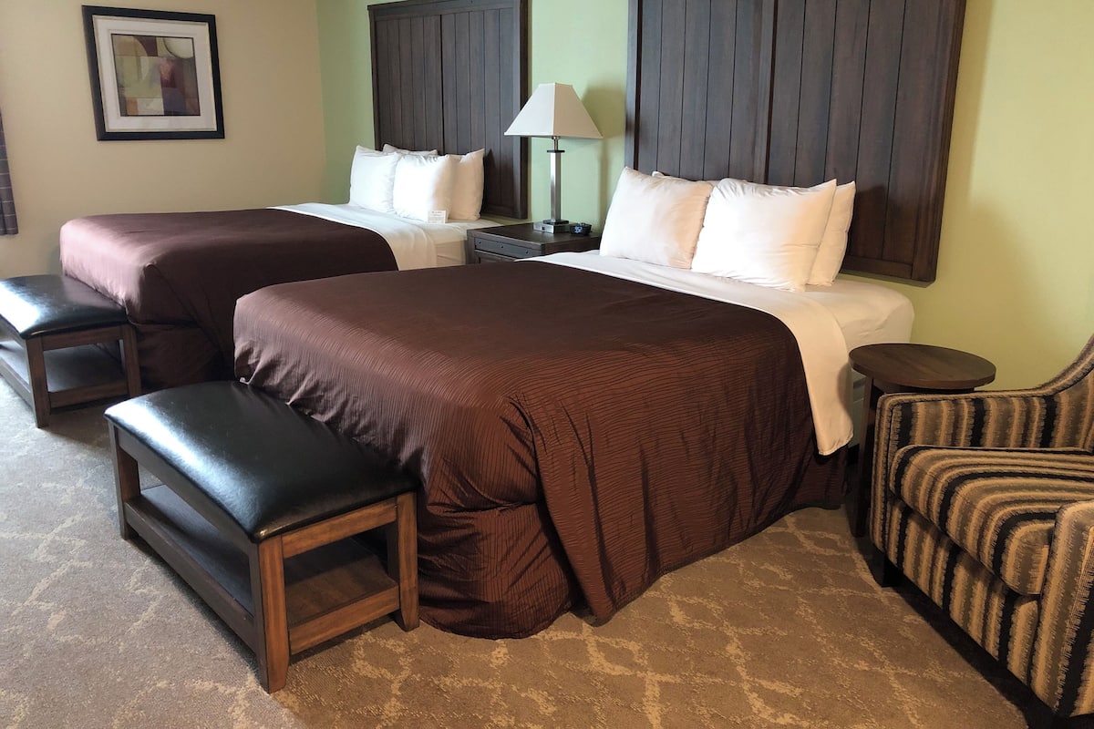 A hotel room with two beds, a bench, a chair, a nightstand with a lamp, and a framed picture on the wall.
