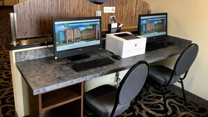 A hotel reception area with two computer workstations, a printer, and a large framed picture of a coastal landscape.