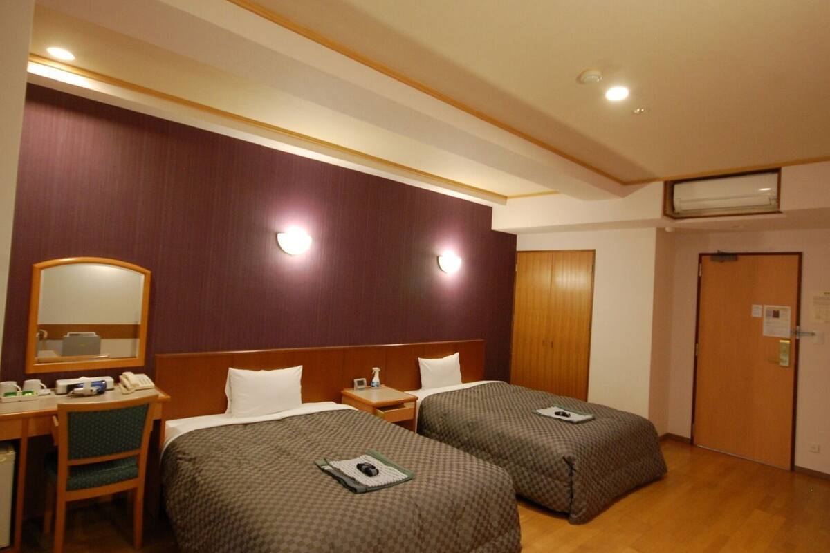 Une chambre d’hôtel avec deux lits, un bureau avec une chaise, un miroir, une armoire en bois et une porte avec un porte-chaussures.