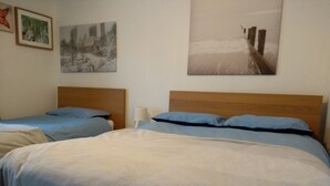 Een hotelkamer met twee bedden, elk met een houten hoofdbord, en een groot bed met een deken van wit en blauw. Er hangen kunstwerken aan de muur, die een stadsgezicht en een strandscène voorstellen.