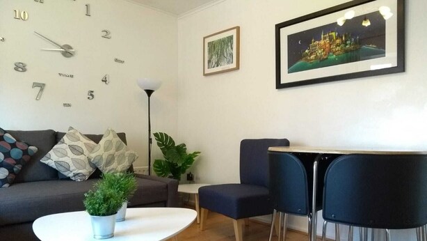 Een moderne woonkamer met een bank, een klein rond tafeltje, twee stoelen, een potplant en een wandklok.