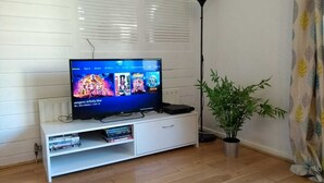Een woonkamer met een flatscreen televisie op een witte televisiemeubel, een potplant en een vloerlamp.