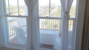 A room with a large window featuring white curtains, a view of a wooden deck with chairs, and a potted plant on the floor.
